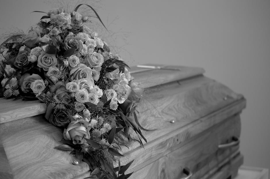 A detailed black-and-white photograph of a large funeral flower spray resting atop a closed wooden casket, featuring an arrangement of densely packed roses and various smaller flowers, with some green