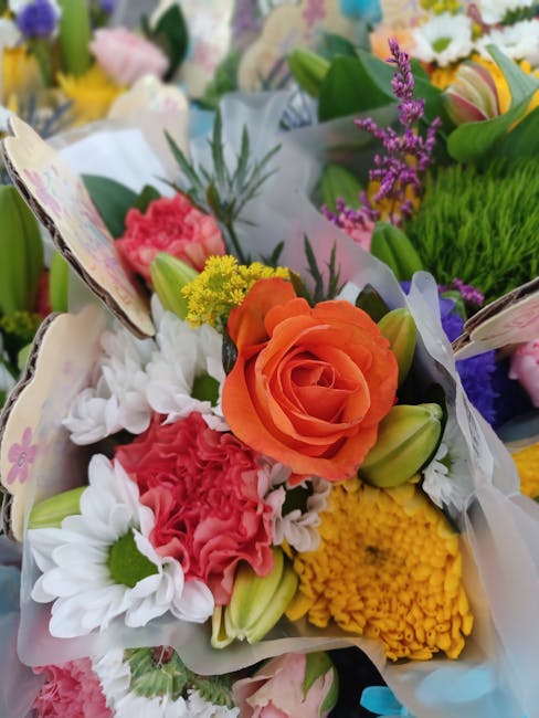 A vibrant floral bouquet featuring a central orange rose surrounded by white daisies, pink carnations, small yellow flowers, and purple lavender sprigs, wrapped in white floral paper with green foliag
