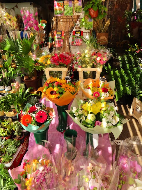 A vibrant display of fresh floral bouquets arranged on a pink cloth-covered table inside a flower shop. The arrangements feature bright, colorful flowers including sunflowers, daisies, gerberas, and r
