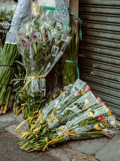 Multiple bouquets of fresh flowers, including pink lilies, yellow tulips, and mixed seasonal blooms, wrapped in clear plastic and tied with yellow and green ribbons, are placed on the pavement outside
