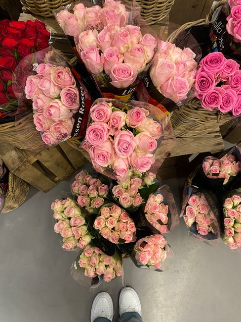 A variety of fresh pink roses in different shades, including pale pink and deeper pink, arranged in multiple bouquets with some wrapped in black paper with white branding and others in clear cellophan